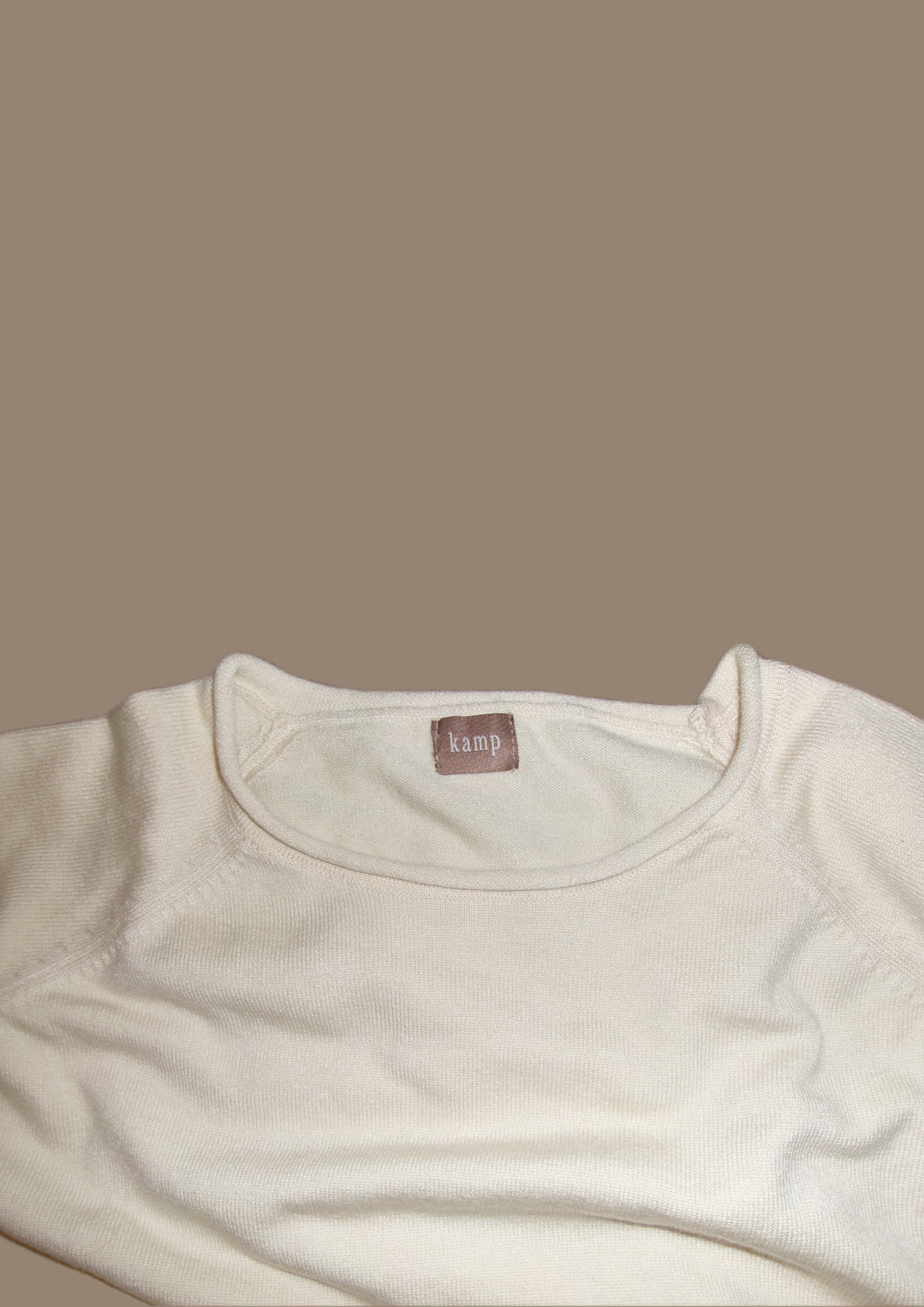 A close-up of a cream-colored long-sleeve top with a crew neck, featuring a small brown brand label that reads 'xiny' at the inside collar, set against a solid taupe background.